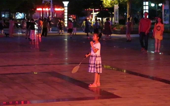 en liten tjej står på ett torg i Kina med ett badminton-racket.