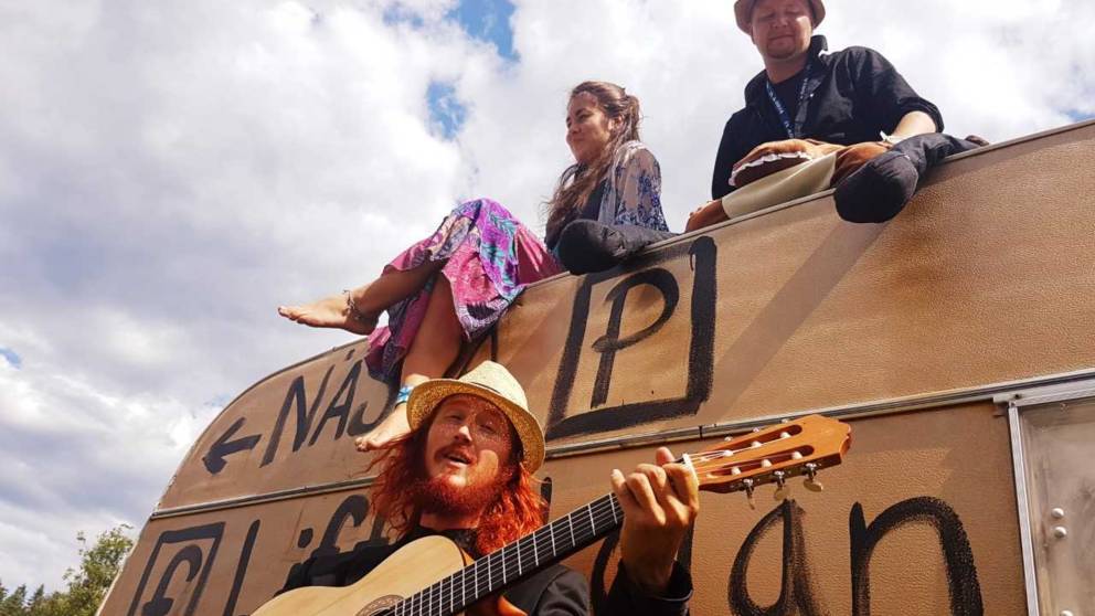En tjej sitter på en husvagn, framför sitter en kille med hatt som spelar gitarr.